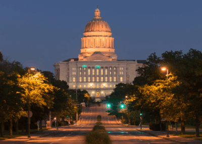 Wide night view of the Missouri State Capitol building in Jefferson City, featuring its illuminated dome and neoclassical facade, with a wide, tree-lined avenue leading up to the entrance.